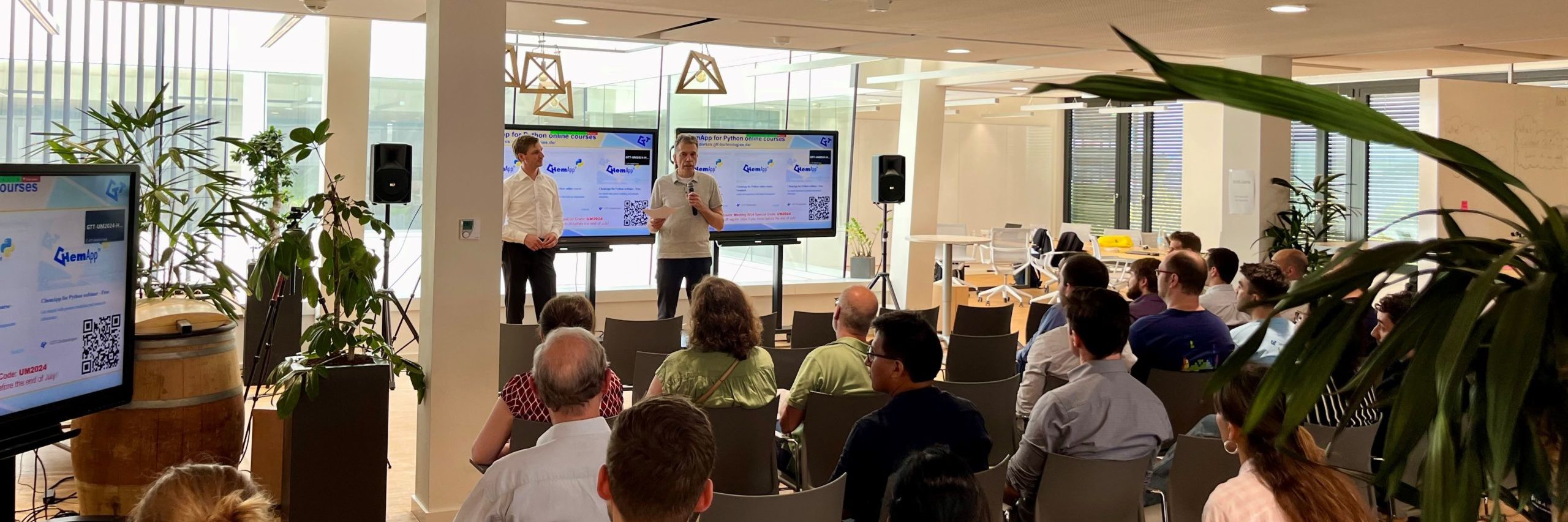 GTT-Technologies' CEOs Moritz to Baben and Stephan Peteren standing in front of a seated audience in a bright, modern conference room with large windows. Two screens display information about courses related to ChemApp for Python. Plants and barrels add decorative touches to the room.