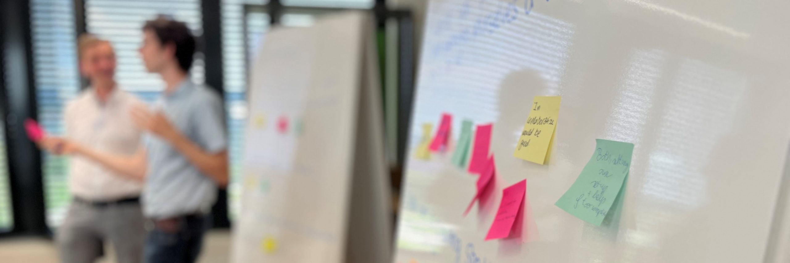 A brainstorming session is taking place in a modern office with large windows. In the foreground, a whiteboard covered with colorful sticky notes in pink, yellow, and green is in focus, featuring handwritten ideas and suggestions. Some notes contain phrases like "In collaboration would be good" and "Both actions are very helpful." In the blurred background, two people are engaged in a discussion, one holding a pink sticky note, emphasizing a collaborative and creative atmosphere. This image serves as the hero image for the GTT-Technologies Newsletter, representing innovation, teamwork, and knowledge sharing.