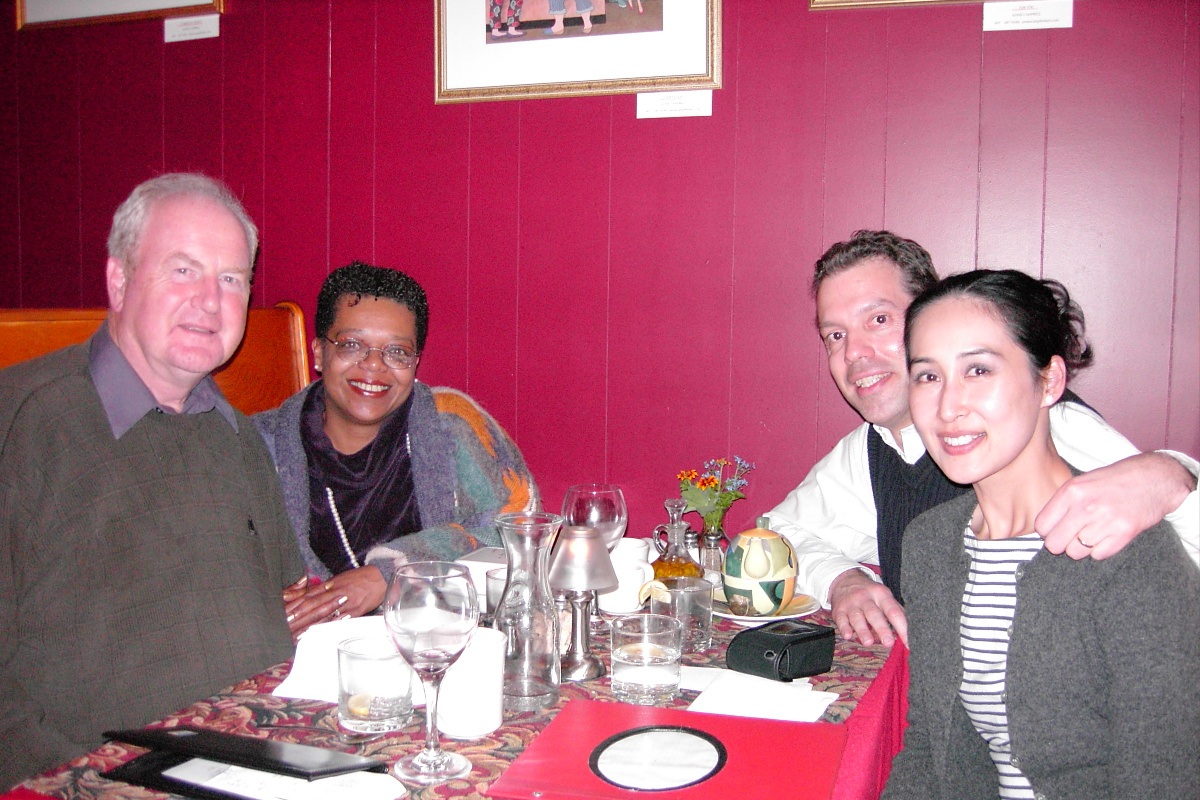 A group of four adults sitting together at a restaurant table during a workshop event. They are smiling toward the camera, with empty glasses, menus, and table decorations in front of them, against a deep red wall with framed artwork.